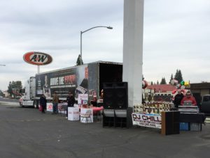 Image of DJ booth and table with trophies and awards set up next to donation boxes in front of white tractor with Drive the Guard themed trailer.