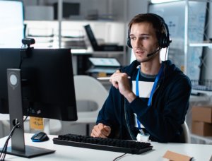 An IT support technician wearing a headset and a blue lanyard sits at a computer and talks with someone on a call. He holds a pen in a workspace with shelves of electronics and equipment behind him.