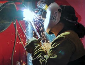A welder in a protective helmet and gloves welding a metal pipe, with bright sparks flying in a shop.