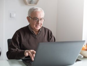 A smiling older man with white hair and glasses, wearing a brown sweater, uses a laptop at a kitchen table at home.